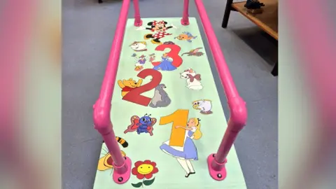 A walking mat with pink parallel metal bars, used for young children with mobility issues to practice walking. The mat is decorated with cartoon characters such as Alice in Wonderland, Minnie Mouse and Winnie the Pooh. 