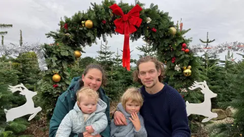 A mother and father sit in front of a giant wreath with their two young children. There is a red bow at the top of the wreath and jumping cut out reindeer on the sides with Christmas trees in the background and baubles and lights on the wreath 