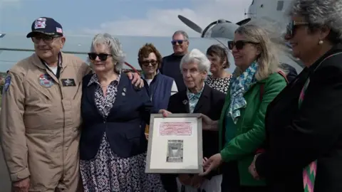 Group photo with a man in a pilot uniform on the left. Two ladies with grey hair and navy blazers hold on to a frame containing a red crayon rubbing and photo.