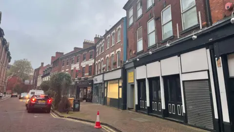 The building subject to the application in the foreground and several other businesses visible at the end of the street.