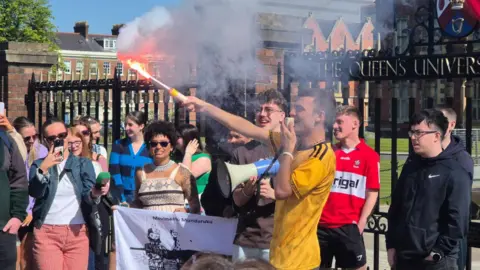 An Cumann Gaelach Eoghan Ó Conghaile, with short black hair, is wearing an amber-coloured sports top and is holding a red flare while speaking into a megaphone. He is surrounded by other students in front of the black iron gates of QUB at a protest calling for bilingual signage.