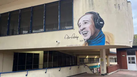 A mural of a fictional radio DJ is painted on the side of a cream-coloured building with blue window frames, with steps shown below a cantilevered section of the building.