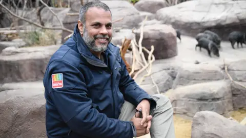 Maarten van der Geest Arun Idoe is sitting on some rocks with his glasses clasped in his hands. He is wearig a jacket with the Bergers' Zoo logo on the shoulder. There are warthogs in the background.