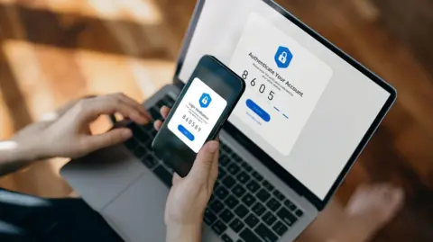 A person holds a smartphone over a laptop to complete a two-factor authentication process. Both screens display security prompts and matching verification codes.