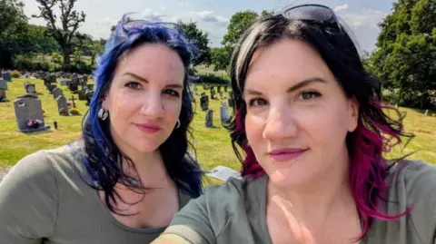 Two women with black, blue and pink hair wearing green tshirts and are stood in a cemetery with green grass and grey head stones. 