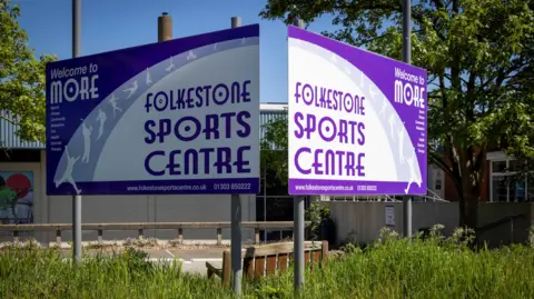 The Sports Trust A purple and white sign reading "FOLKESTONE SPORTS CENTRE" sat in front of a building.