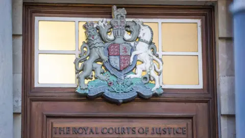 A court crest on a window above a brown wooden door which says 'the royal courts of justice'. The crest is silver with green, grey, blue and red colours on it. The wall surround the crest and door is stone.
