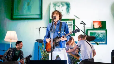 Paul Campbell Razorlight on stage at Belladrum. Lead singer Johnny Borrell, wearing a blue denim shirt and white jeans, stands at a microphone singing while playing a guitar.