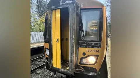 West Midlands Railway A yellow train is pictured at a train station with damage to its front. The coupling section appears to have missing parts with some debris inside the train. 