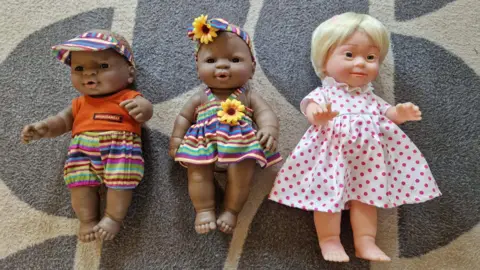 St John's preschool Three dolls lying on a carpet. Two of the dolls are black and another doll is white and has Down syndrome. 