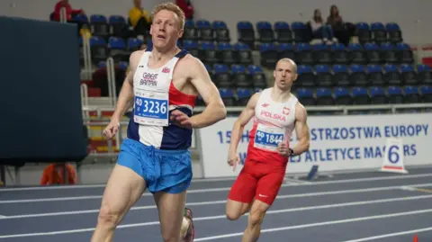 supplied Two men sprint around a blue athletics track