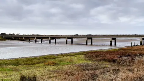 David Dixon/Geograph the Shard bridge over the river Wyre, a long bridge spanning across the wide, calm river with wild grasses to the foreground