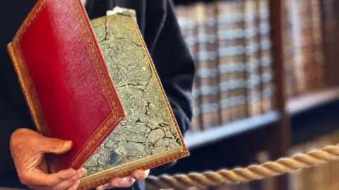 Ushaw Historic House A man holds a half open book - the Nuremberg Chronicle, dating from 1493. It has a red cover with gold printed edging, and the front page is marbled paper. All that can be seen of the man holding it are his hands and arms encased in a dark jacket. Behind, and out of focus, is a rope barrier.
