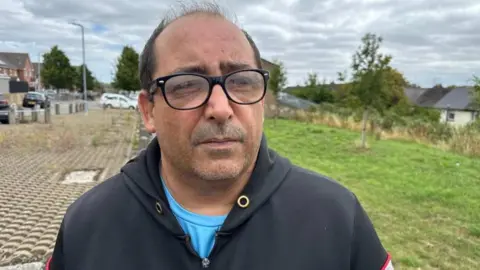 BBC A man in a black hooded top, with a blue t-shirt underneath with black glasses. It is a head and shoulders shot of him.