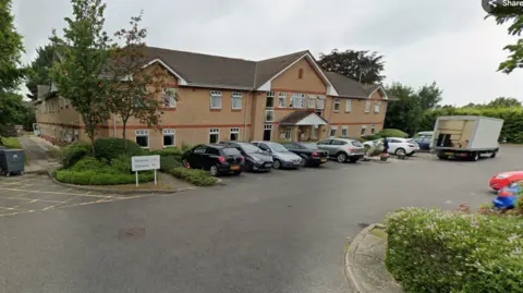 Google The outside of the care home, which has several cars parked in the car park. It is a two storey modern brick building, probably 1990s, with pairs of small windows all around the building. There are trees and bushes surrounding the home.