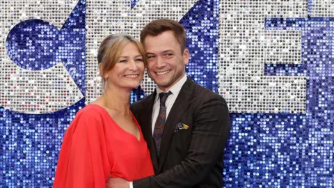 Tristan Fewings/Getty Images An older blonde woman in a red dress and Egerton with brown hair and wearing a suit. They're on a blue "red carpet" and it has a blue sequin background with silver sequin words behind them. 