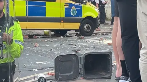 A wheelie bin and litter is seen all over a street in Southport. There is a police van in the background and a police officer with a riot shield to the side of the picture.