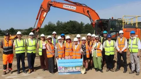 People wearing hard hats and high-visibility vests stand at a housing development site in front of an excavator, holding a sign promoting a council housing project.