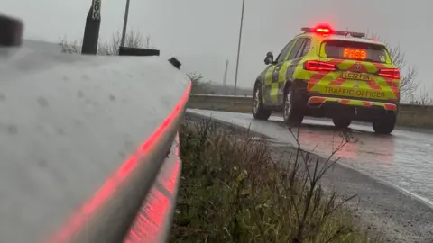 A traffic officer car parked with its red lights illuminated, with the lights reflecting off a metal side barrier beside the road.