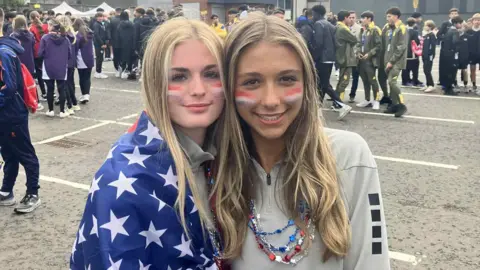 BBC Two girls, one draped in an American flag, and the other in a grey sweatshirt are standing in a car park. They are both smiling.