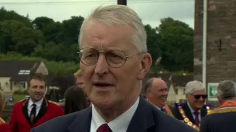 Hilary Benn, dressed in a suit, stands in front of a number of members of the Orange Order