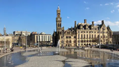 A wide shot of the outside of Bradford City Hall and City Park in front of it with water fountains spraying.