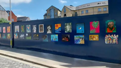 Multiple bright artworks on dark blue hoardings, the sky in the background is blue. 