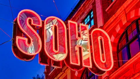 Getty Images Large red Soho sign with the outline of the letters bordered by small light bulbs. It is a sign attached to a building and it is evening, with dark blue sky in the background.