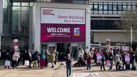 A crowd of people are stood outside Sheffield Hallam University with purple and white strike placards