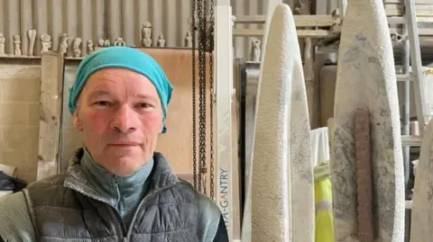 Tom Percival/BBC Nicolas Moreton is looking at the camera. He is wearing a turquoise head covering and a blue top and black gilet jacket. He is standing in a studio and there are stone sculptures behind him. 