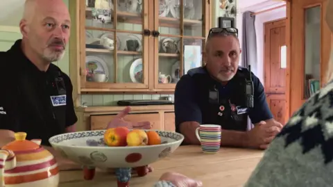 BBC A Trading Standards officer and a police community support officer sit at a kitchen table. There is a fruit bowl and a woman's grey jumper is visible.