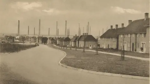 Forterra A sepia photograph showing one of the main roads in Stewartby. In the foreground is the road, and newly planted trees. Behind them a series of brick houses all with the same style of white framed windows. Behind those houses, you can see the incredibly tall chimneys of Stewartby brickworks.