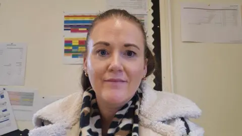 Aisling has brown hair tied up and is smiling at the camera. She has gold earrings on and a leopard print scarf and teddy bear woolly white coat on. 
