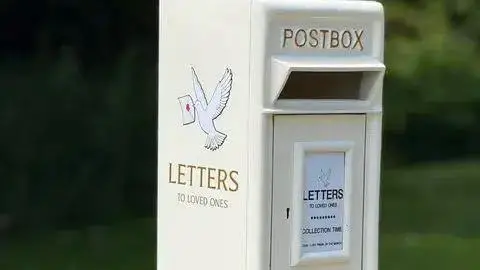 A white post box with the words "Letters to loved ones" on the side below a dove flying with a letter in its mouth. It has the traditional Post Box writing on the front with a little white plaque on the front.