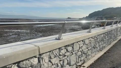 Part of the finished new wall along the promenade in Mumbles, with sun glinting off the metal handrail, and the beach, sea and pier in the background