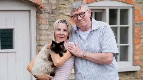 Postcode Lottery A couple stand in front of a stone cottage. They are holding a brown and cream coloured cat. The woman on the left of the image has blonde hair and is wearing a striped top. The man on the right of the image is wearing a blue shirt and has grey hair and glasses.