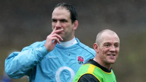 David Rogers/Getty Images Mike Tindall walks away while talking to Martin Johnson during England Rugby Union squad training. Martin is tall with dark hair and a blue training shirt on, and has a finger resting on his nostril. While Mike Tindall is bald, has his mouth open in a smile, and is wearing a black T-shirt and a green and yellow vest.