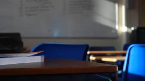 A classroom with a whiteboard, desks and blue chairs. The whiteboard has blurred black writing on it. Sunlight is shining on the whiteboard. There is a book on a table.
