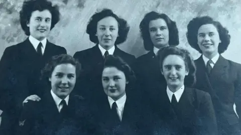 Barbara Quevâtre A formal black and white photo of seven women in two rows, four at the back and three at the front. All smiling, with dark hair, smartly dressed in their uniform of white shirt with dark coloured tie and jacket.