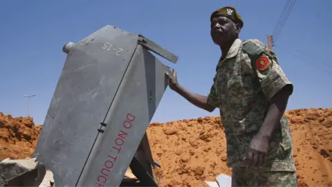A man in uniform with a red beret stands in a hole filled with drone components in El Obeid, Sudan - March 2026