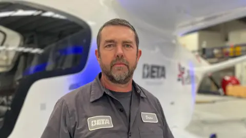 Brian Jenkins stood in front of the aircraft. He's wearing a dark coloured shirt which says "Beta" on one side and "Brian" on the other. 