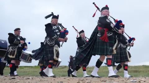 PA Media Scottish pipers walk along beach. They wear kilts and carry bagpipes and drums