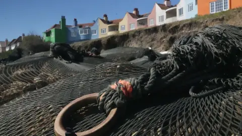 John Fairhall/BBC A close-up of a black net holding rocks as part of a sea defences. Beyond it is a shallow sandy cliff and above that a block of flats painted in vibrant colours. 