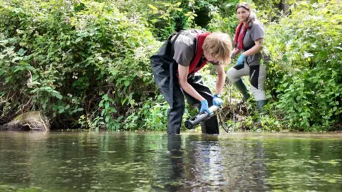 A man wearing a life jacket and waders stands in the River Test preparing to place a water quality monitor on the riverbed