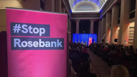 Stop Rosebank campaign event in Edinburgh. An audience of people sitting in chairs looks towards a stage. Four people - who are too far away to be identified - are having a discussion. There is a pink sign in the foreground, saying #Stop Rosebank 