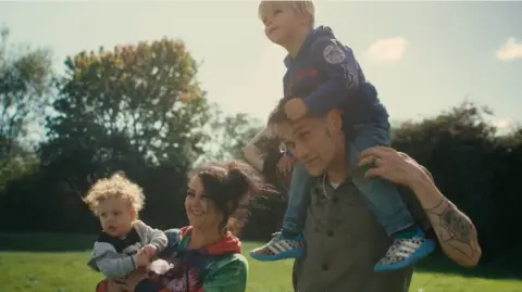 Isaac James Rob with his family. He is wearing a green shirt and has a child sat on his shoulders. His partner is stood next to him holding their other child in her arms.