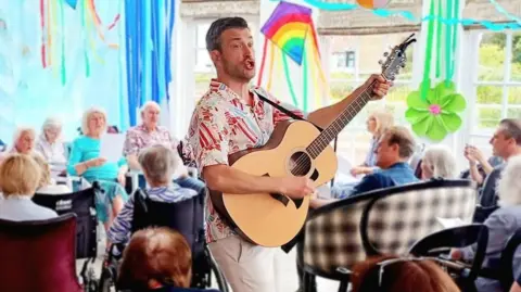 supplied A young man holding a guitar and smiling whilst elderly people in wheelchairs look on.