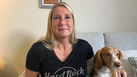 A blond woman wearing a V-neck black top, sits on a sofa with her arm around a chestnut coloured Cocker Spaniel. She is smiling into the camera.