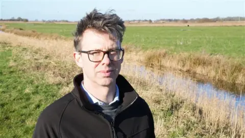 John Fairhall/BBC Eliot Lyne is looking at the camera and is stood in a field. He is wearing glasses and is wearing a black sweatshirt, shirt and grey jumper. 