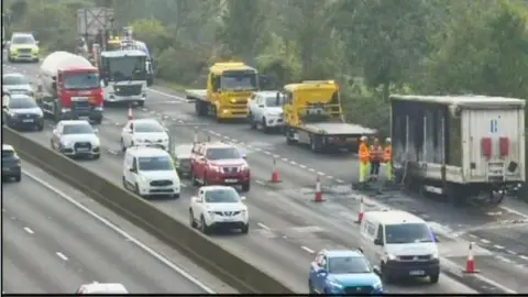 National Highways A CCTV image showing cars passing by the burning cargo via two lanes. The burning load is cordoned off with three people in high vis standing by.